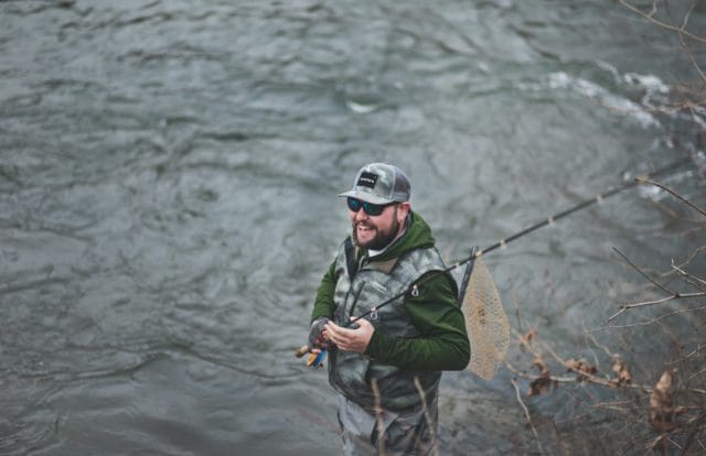 man smiling holding simms black fishing rod near body of water